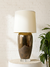 Load image into Gallery viewer, Image 1 of 17: Vintage table lamp featuring a tall rounded body with a reflective dark metallic upper finish, textured charcoal lower section, clear Lucite-style circular base, and illuminated ivory cylindrical drum shade.
