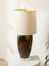 Load image into Gallery viewer, Image 3 of 17: Vintage table lamp featuring a tall rounded body with a reflective dark metallic upper finish, textured charcoal lower section, clear Lucite-style circular base, and illuminated ivory cylindrical drum shade.

