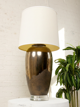 Load image into Gallery viewer, Image 7 of 17: Vintage table lamp featuring a tall rounded body with a reflective dark metallic upper finish, textured charcoal lower section, clear Lucite-style circular base, and illuminated ivory cylindrical drum shade.
