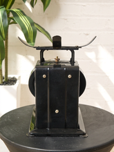 Load image into Gallery viewer, Image 6 of 10: A vintage black metal coffee grinder with brass screws and a curved handle, displayed on a black round table against a white brick wall with a green plant in the background.
