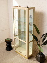 Load image into Gallery viewer, Image 6 of 24: A cream-colored mid-century modern glass display cabinet with multiple shelves, shown in a styled room corner with a white brick wall, a black side table, and a potted plant.
