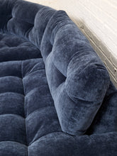 Load image into Gallery viewer, Image 10 of 16: Close-up of a tufted velvet sofa in a dark blue-gray color, showing the rich fabric texture and button tufting details against a white brick wall background.
