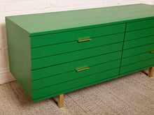 Load image into Gallery viewer, Image 6 of 15: A mid-century modern green dresser with six drawers, gold-toned metal handles, and light wood legs, positioned against a white brick wall on a textured beige rug.
