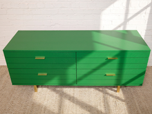 Load image into Gallery viewer, Image 5 of 15: A bright green mid-century modern sideboard with gold-tone handles and legs, set against a white brick wall with natural window shadows.
