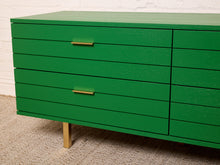 Load image into Gallery viewer, Image 7 of 15: A bright green wooden sideboard or dresser with six drawers, featuring slim gold metal handles and a single gold leg, set against a white brick wall and textured carpet.
