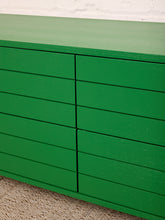 Load image into Gallery viewer, Image 11 of 15: A close-up of a bright green wooden dresser with multiple drawers, positioned against a white brick wall and a textured beige carpet.
