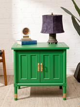 Load image into Gallery viewer, Image 4 of 21: A bright green wooden side table or cabinet with two paneled doors and gold-tone handles, styled in a room setting with a dark purple lamp, a small clock, and books on top, set against a white brick wall.
