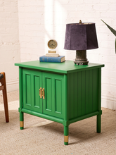 Load image into Gallery viewer, Image 5 of 21: A bright green wooden nightstand with two cabinet doors, gold-tone handles, and gold-tone caps on the legs, styled in a room setting with a purple lamp, books, and a clock on top.
