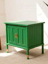 Load image into Gallery viewer, Image 8 of 21: A bright green wooden nightstand with double doors, gold-tone handles, and gold-colored caps on the legs, set on a beige carpet against a white brick wall.
