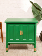 Load image into Gallery viewer, Image 7 of 21: A bright green two-door cabinet with gold handles and gold-colored feet, set against a white brick wall background.
