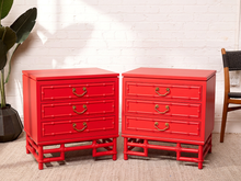 Load image into Gallery viewer, Image 1 of 21: Pair of vintage 1960s Hollywood Regency nightstands featuring vibrant poppy red painted finishes, three drawers each with brass-tone curved handles, decorative faux bamboo trim, and sculptural bamboo-inspired bases.
