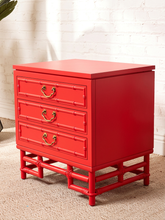 Load image into Gallery viewer, Image 8 of 21: A bright red three-drawer nightstand with a bamboo-inspired frame design and brass handles, set against a white brick wall and beige carpet.
