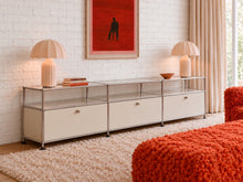 Load image into Gallery viewer, Modern three-drawer media console with chrome-toned frame, glass shelves, light neutral cabinet fronts, and open storage compartments.
