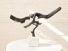 Load image into Gallery viewer, Abstract black metal sculpture on a white marble base, displayed on a white round pedestal against a white brick wall with soft sunlight and shadows.
