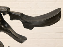Load image into Gallery viewer, Close-up of a dark charcoal-colored abstract metal sculpture against a light beige brick wall background.
