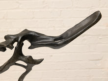 Load image into Gallery viewer, Abstract organic-shaped dark metal sculpture against a light beige brick wall background.

