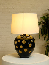 Load image into Gallery viewer, A table lamp with a black ceramic base featuring gold circular cutouts and a warm white fabric drum shade, displayed on a white pedestal against a white brick wall with a green plant in the background.
