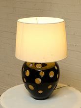Load image into Gallery viewer, A table lamp with a glowing warm white fabric drum shade and a glossy black ceramic base featuring circular cutouts, set on a white round table against a white brick wall.
