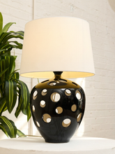 Load image into Gallery viewer, A table lamp with a glossy black ceramic base featuring circular cutouts and a warm white fabric drum shade, set on a white surface next to a green plant against a white brick wall.
