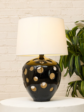 Load image into Gallery viewer, A table lamp with a glossy black ceramic base featuring circular cutouts and a warm gold-toned collar, topped with a large off-white drum shade. It sits on a white round pedestal against a white brick wall, with a green plant visible on the right.
