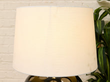 Load image into Gallery viewer, A close-up of a table lamp with a large, warm off-white fabric drum shade, set against a white painted brick wall with a green plant visible on the right.
