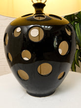 Load image into Gallery viewer, A glossy black ceramic table lamp with a lid and multiple circular cutouts, shown on a white surface with a beige lampshade and a green plant in the background.
