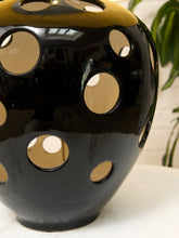 Load image into Gallery viewer, A glossy black ceramic vase with a gold-toned glazed top and multiple circular cutouts, shown close up on a white surface with a blurred green plant in the background.
