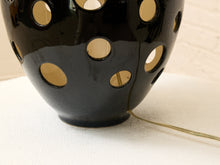 Load image into Gallery viewer, A close-up of a glossy black ceramic lamp base with circular cutouts, showing a warm yellow light glowing inside, set on a white textured fabric surface.
