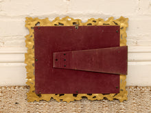Load image into Gallery viewer, The back of a rectangular picture frame with a scalloped gold metal border and a dark red backplate, resting on a woven rug against a white brick wall.
