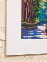Load image into Gallery viewer, A close-up of a framed art print featuring a colorful landscape painting of a redwood forest path, mounted on a textured off-white brick wall.

