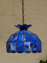 Load image into Gallery viewer, A vintage-style stained glass pendant light with a cobalt blue glass shade featuring leaf patterns and dark metal framing, hanging against a white brick wall.
