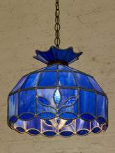 Load image into Gallery viewer, A stained glass pendant light with a vibrant cobalt blue glass shade featuring a leaf motif and a scalloped lower edge, suspended by a dark metal chain against a textured beige brick wall.
