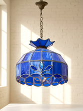 Load image into Gallery viewer, A stained glass pendant light with a vibrant blue glass shade featuring a floral leaf pattern and a scalloped bottom edge, suspended by a dark metal chain in front of a white brick wall with soft window light.

