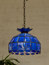 Load image into Gallery viewer, A stained glass pendant light with a cobalt blue glass shade and dark metal frame, hanging against a white brick wall.
