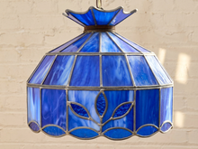 Load image into Gallery viewer, A stained glass pendant light with a blue and white marbled pattern, featuring a leaf motif on the lower panel and a scalloped glass top, suspended against a light beige brick wall.
