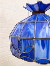Load image into Gallery viewer, A close-up of a hanging pendant light with a cobalt blue stained-glass shade and dark metal frame, set against a white brick wall.
