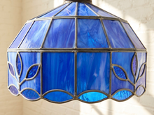 Load image into Gallery viewer, A close-up of a stained glass pendant light shade with vibrant blue glass panels and dark metal leaf-shaped outlines, shown against a softly blurred light background.
