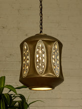 Load image into Gallery viewer, A hanging pendant light with a bronze-toned metal body, three oval cutout panels with warm glowing bulbs inside, and a dark chain, shown against a white brick wall with green leaves at the lower left.
