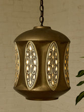 Load image into Gallery viewer, A vintage-style brass pendant light with ornate cutout panels, glowing with warm light against a white brick wall background.
