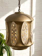 Load image into Gallery viewer, A hanging brass pendant light with ornate cutout panels, shown illuminated against a white brick wall with a plant leaf visible at the lower left.
