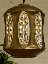 Load image into Gallery viewer, A hanging pendant light with a bronze-finished metal body and three vertical oval cutout panels that glow warm yellow from within, shown against a light brick wall with a small plant leaf at the lower left.
