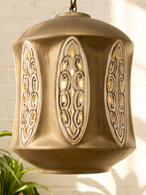 Load image into Gallery viewer, A hanging pendant light with a warm metallic bronze finish and ornate cutout panels, shown against a white brick wall with soft sunlight and a small plant visible at the lower left.
