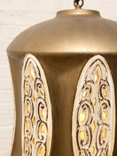 Load image into Gallery viewer, Close-up of a hanging lantern with a distressed metallic gold finish and two arched cutout panels showing warm yellow light inside, set against a white brick wall.
