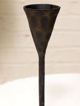 Load image into Gallery viewer, A dark, rustic metal candle holder with a textured, hammered finish, featuring a wide conical cup and a long straight stem, shown against a neutral light beige background.
