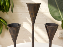 Load image into Gallery viewer, Three rustic dark metal cone-shaped candle holders with long stems, displayed on a white surface against a white brick wall with green plants in the background.

