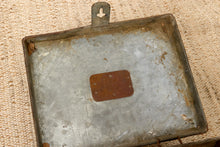 Load image into Gallery viewer, A vintage rectangular metal tray with a hinged top handle, showing signs of rust and age, placed on a textured beige woven rug.
