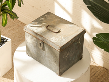 Load image into Gallery viewer, A distressed vintage metal storage chest with a hinged lid and latch, displayed on a white pedestal in a sunlit room with plants.
