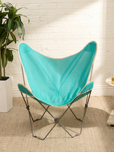 Load image into Gallery viewer, A turquoise butterfly-style chair with a thin metal frame, set on a beige textured rug against a white brick wall with a potted plant to the left.
