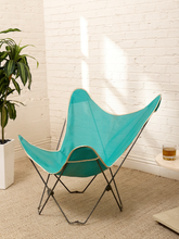 Load image into Gallery viewer, A butterfly-style chair with a bright turquoise mesh seat and back, supported by a black metal frame, set in a room with a white brick wall, beige carpet, a potted plant, and a small side table with a drink.
