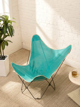 Load image into Gallery viewer, A butterfly-style chair with a bright turquoise seat and back, supported by a thin black metal frame, styled in a sunlit room with a white brick wall, beige rug, and a potted plant.
